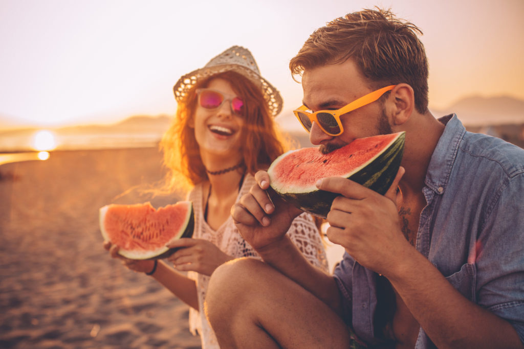 Juicy and sweet summer Hipster couple sitting on the beach and eating watermelon. Wearing casual summer clothing, hat and sunglasses. Enjoying in sunset by the sea.