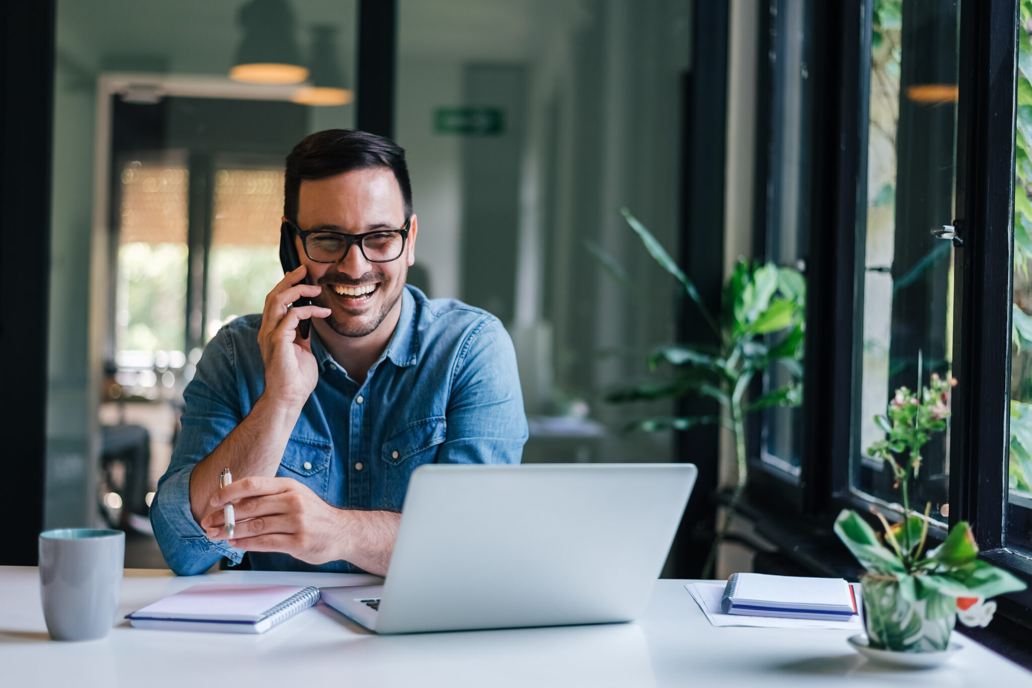 Portrait of young smiling cheerful entrepreneur in casual office making phone call while working with laptop Portrait of young smiling cheerful entrepreneur in casual office making phone call while working with laptop